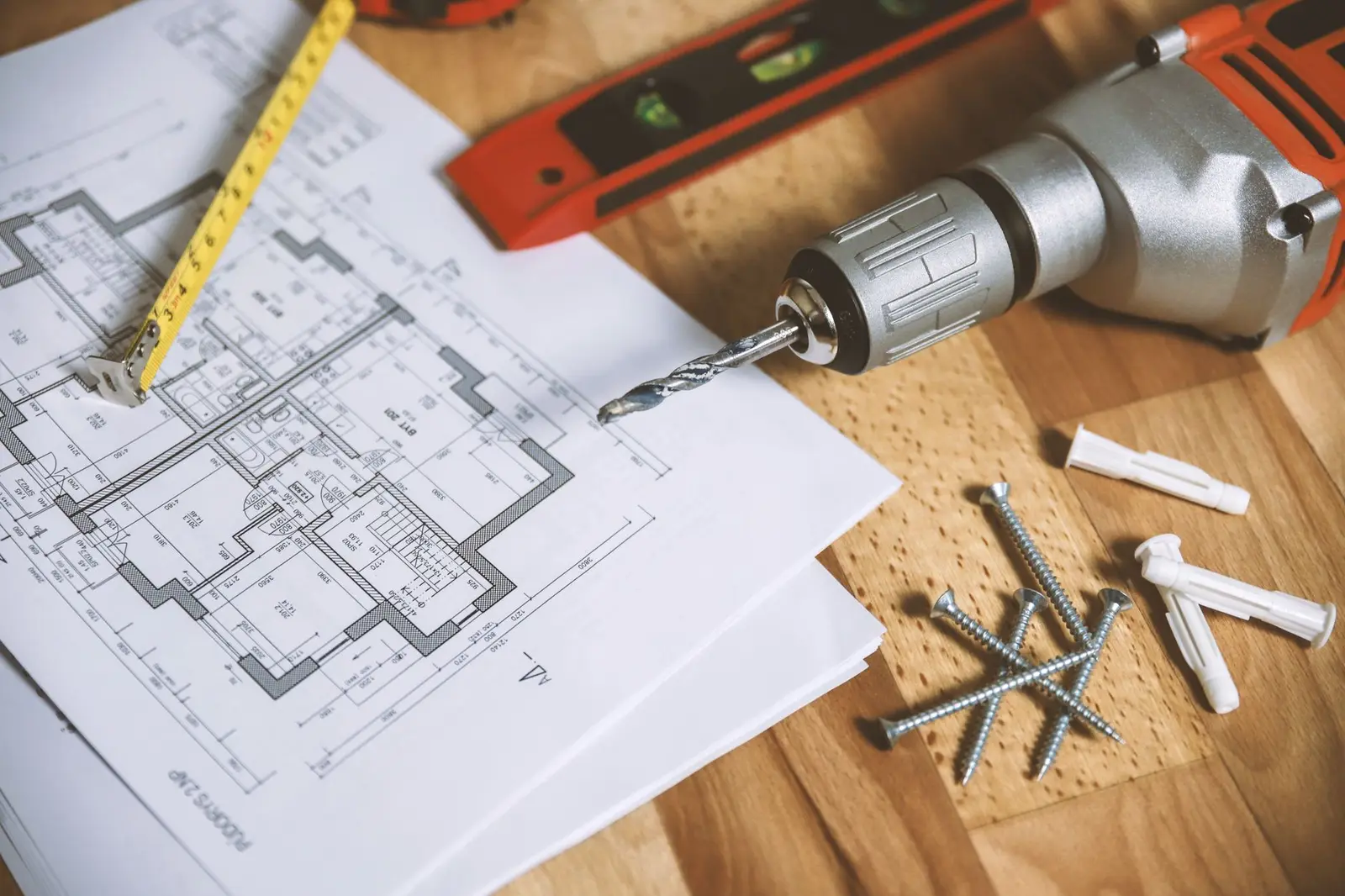 Plans d’architecte, mètre ruban jaune et perceuse posés sur une table en bois avec des chevilles et des vis.